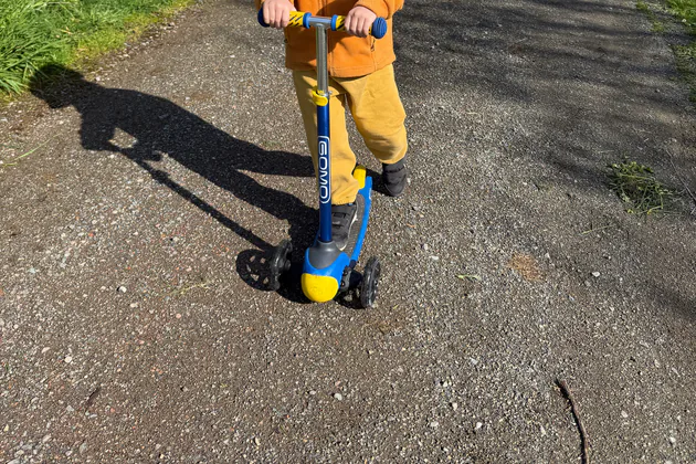 Child holding the adjustable bar of a blue and yellow GOMO three-wheel scooter on dry pavement