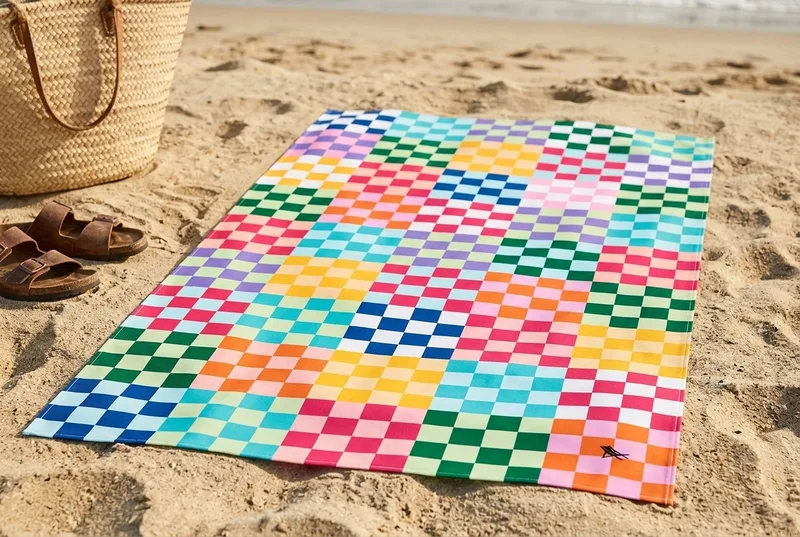 Dock & Bay Kid's quick-dry towel in a multicolour checkerboard print, spread flat on dry beach sand beside a straw bag and leather sandals