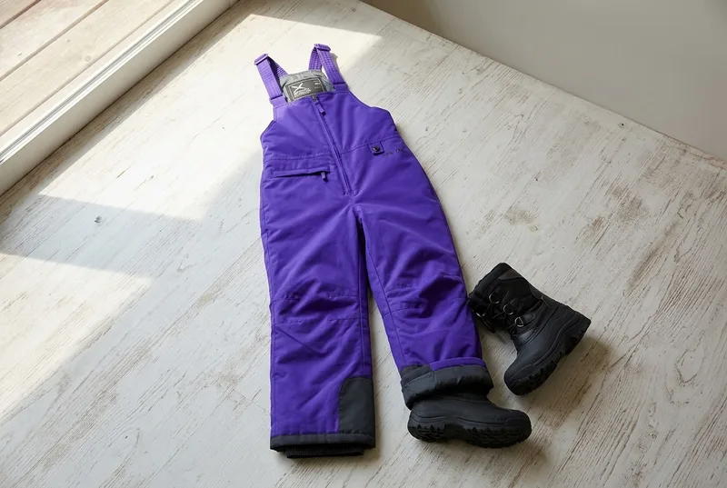 Purple Arctix Kids Insulated Snow Bib laid beside black winter boots on a pale wooden floor