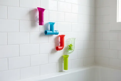 Five Boon Pipes bath toys in magenta, blue, red, green and clear plastic suction-mounted on white subway tile above a bathtub, arranged so water runs from one into the next