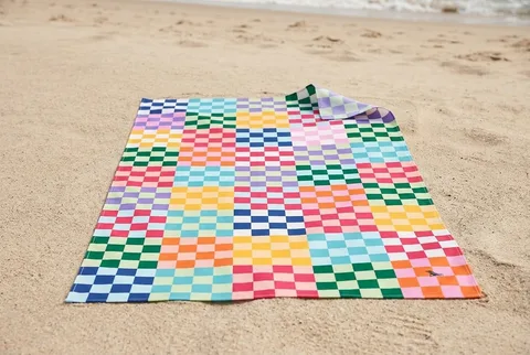 Dock & Bay Kid's quick-dry towel in a multicolour checkerboard print, spread flat on dry beach sand with one corner folded back