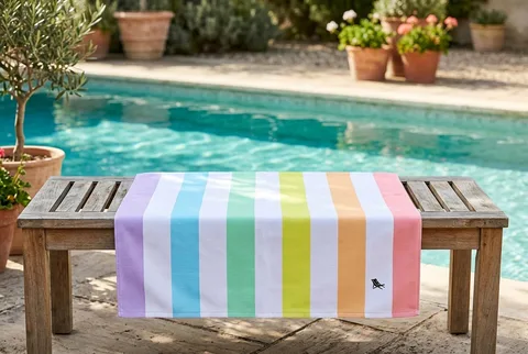 Dock & Bay large quick-dry towel in pastel rainbow stripes, draped over a slatted wooden bench beside a pool