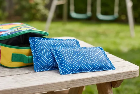 Two Fit & Fresh Cool Coolers ice packs in a blue printed pattern on a weathered picnic table beside an open insulated lunch bag