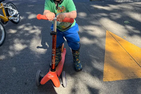 Child holding the adjustable bar of an orange Mini Micro Deluxe scooter beside a splash pad