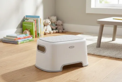 Gray and white OXO Tot step stool on a wooden floor near picture books and stuffed animals