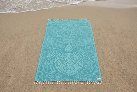 Sand Cloud Turkish beach towel in teal with a woven sea-turtle motif and tasselled ends, spread flat on damp beach sand near the tide line