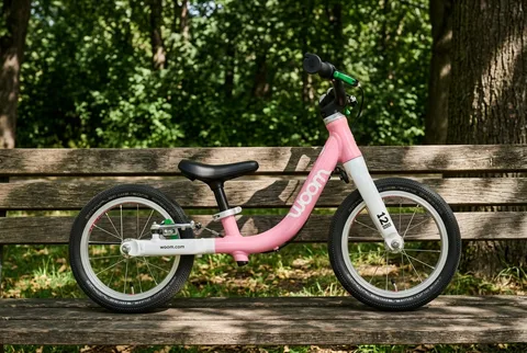 Pink Woom GO 1 balance bike leaning on a wooden park bench with trees behind